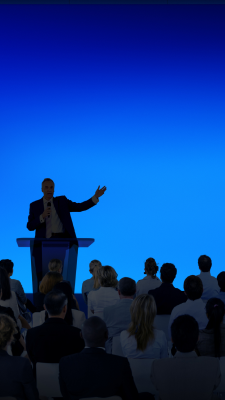A man speaking to a crowd from a podium, against a blue screen 